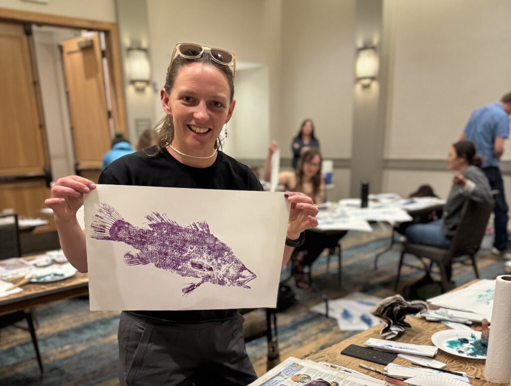 A woman holds up a gyotaku print in purple paint of a rockfish