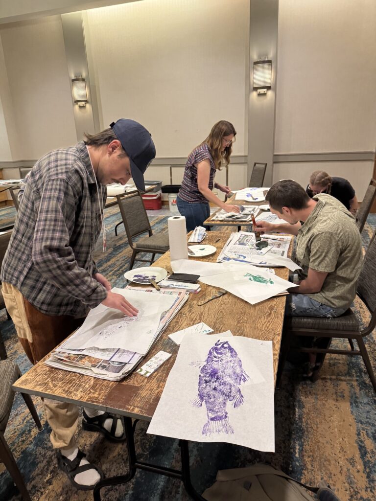 Several people sitting or standing at a table participating in a gyotaku painting project. One is actively adding paint to a fish, while two are pressing paper on top of already painted. A finished print in purple paint sits on the table closest to the camera.